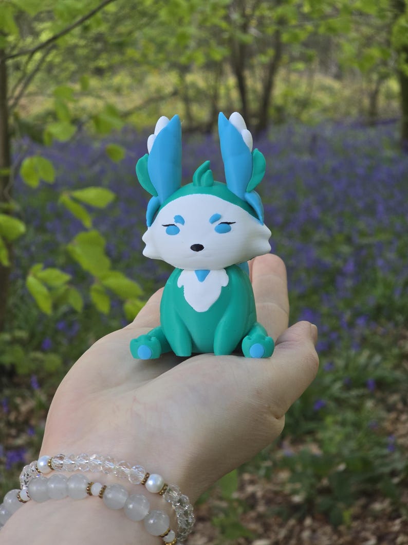 A close-up view of the Fall Flutterfox 3D-printed miniature, sitting on a hand, against a backdrop of natural forest and bluebells. The figure has a rich turquoise and dark red color scheme.