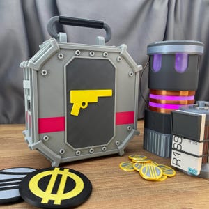 Op de afbeelding: Een grijze, doosvormige container met een geel pistoolembleem, een zwarte handgreep en roze accenten. Daarnaast een cilindrisch apparaat met paarse en roze lichten, munten en een stapel blokken. De items lijken rekwisieten te zijn.