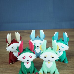 A product group photograph on a light wooden table against a grey-blue background. It features all five Palia Flutterfox 3D-printed miniature variants sitting in a clustered formation. From left to right: Autumn (dark red), Summer (teal with pink), Winter (light blue), Spring (mint green), and Leafy (teal with blue).