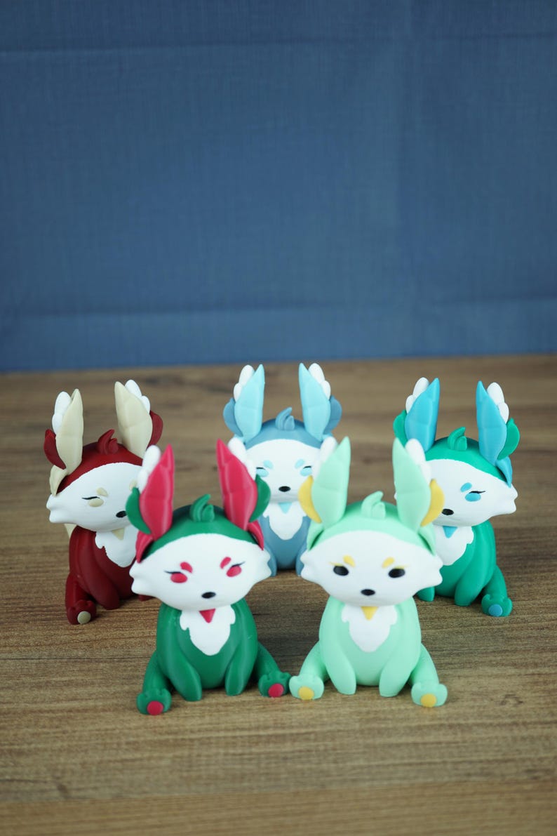 A product group photograph on a light wooden table against a grey-blue background. It features all five Palia Flutterfox 3D-printed miniature variants sitting in a clustered formation. From left to right: Autumn (dark red), Summer (teal with pink), Winter (light blue), Spring (mint green), and Leafy (teal with blue).