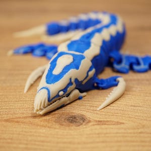 Könnte beinhalten: Ein 3D-gedrucktes Modell eines Plesiosauriers in Blau und Off-White. Das Wasserreptil hat einen langen Hals, Flossen und einen detaillierten Kopf. Das Modell liegt auf einer Holzoberfläche und zeigt sein detailliertes Design und den Farbkontrast.