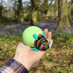 A lifestyle photo showing a person holding the Palia Lantern Bug collectible in their palm to demonstrate its 11cm scale. The person is wearing a blue flannel shirt, and the background is a stunning, sun-drenched forest filled with blooming purple bluebells. The natural sunlight highlights the 3D printed details and the vibrant green, brown, and turquoise colors of the bug. It provides a clear sense of size and shows how the piece looks as a high-quality physical item for collectors.