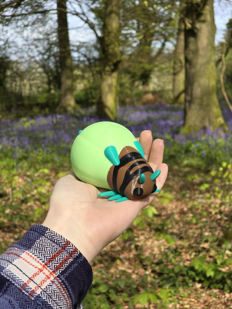 A lifestyle photo showing a person holding the Palia Lantern Bug collectible in their palm to demonstrate its 11cm scale. The person is wearing a blue flannel shirt, and the background is a stunning, sun-drenched forest filled with blooming purple bluebells. The natural sunlight highlights the 3D printed details and the vibrant green, brown, and turquoise colors of the bug. It provides a clear sense of size and shows how the piece looks as a high-quality physical item for collectors.