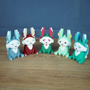 A frontal group product photograph on a light wooden table against a grey-blue background. All five Palia Flutterfox 3D-printed miniature variants—Winter (light blue), Autumn (dark red), Summer (teal with pink), Spring (mint green), and Leafy (teal with blue)—are sitting side-by-side in a curved, evenly-spaced row.