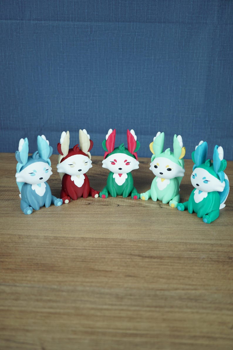 A frontal group product photograph on a light wooden table against a grey-blue background. All five Palia Flutterfox 3D-printed miniature variants—Winter (light blue), Autumn (dark red), Summer (teal with pink), Spring (mint green), and Leafy (teal with blue)—are sitting side-by-side in a curved, evenly-spaced row.