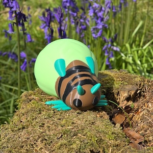 A high-quality hero shot of the 3D printed Palia Lantern Bug figurine resting on a textured, moss-covered tree stump. The vibrant turquoise legs and antennae, brown striped body, and large light-green abdomen pop against the natural forest background. In the soft-focus distance, a field of purple bluebells and green trees creates a magical, "Bahari Bay" atmosphere. This shot perfectly captures the whimsical, game-accurate feel of the collectible in a natural outdoor setting.