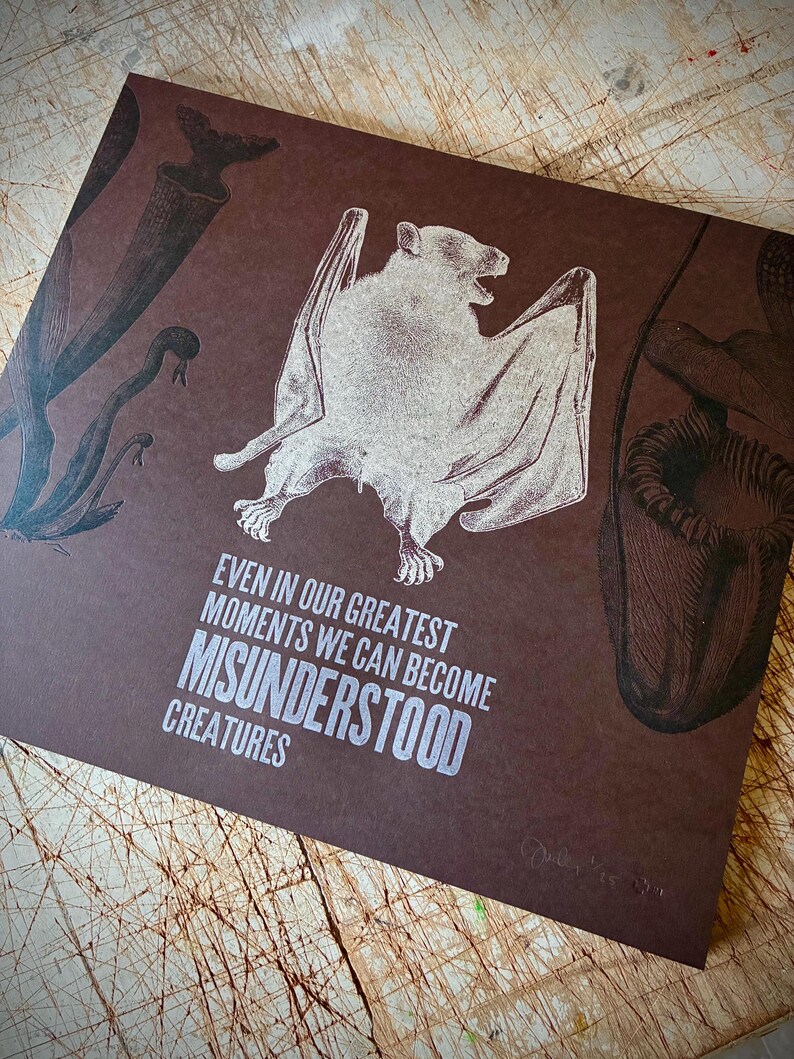 May include: A brown print with a detailed illustration of a bat and botanical drawings. The text reads "Even in our greatest moments we can become misunderstood creatures." The print has a vintage aesthetic.