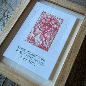 May include: A framed print featuring a red ink illustration of a human brain with text labels, surrounded by embossed raindrop patterns on white paper. Below the illustration, the text reads: YOUR SECRET CODE IS NOT LOST ON ME, I SEE YOU.