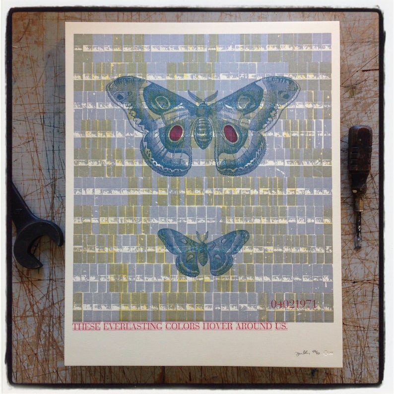 Pu&ograve; includere: Una stampa in stile vintage con due falene blu e verdi con accenti rossi su uno sfondo di quadrati gialli e blu. Il testo "Questi colori eterni aleggiano intorno a noi" &egrave; stampato sotto le falene.