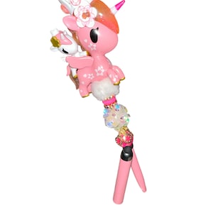Peut inclure: Un stylo surmonté d'une figurine de licorne rose avec une couronne blanche, une plus petite licorne blanche sur le dos et des motifs de fleurs de cerisier. Le corps du stylo est orné de perles irisées, sphériques et roses au-dessus d'une base rose.