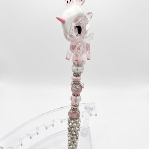 Pode incluir: Uma caneta decorativa com um topo de unicórnio rosa translúcido com duas pequenas figuras de gatos na cabeça. O corpo da caneta é adornado com uma série de contas empilhadas em tons de rosa, prata e transparente iridescente, incluindo espaçadores facetados e metálicos.