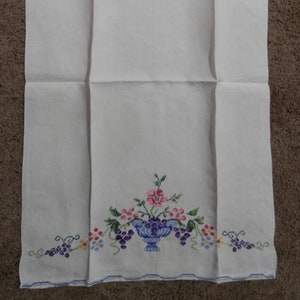 Könnte beinhalten: Weißes Leinenhandtuch mit blauem, gesticktem Rand. Das Handtuch zeigt ein Kreuzstichmuster einer blauen Vase, die mit rosa und lila Blumen und Trauben überquillt. Der untere Rand hat zusätzliche Trauben.