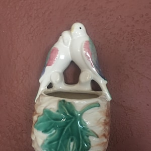 May include: A white ceramic planter with a green leaf design and two white birds perched on the top. The birds are painted with pink, green, and blue accents.