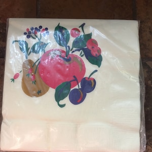 May include: A white paper napkin with a colorful fruit design. The design features a red apple, a brown pear, and purple cherries on a green vine.