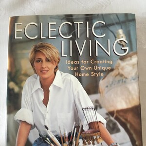 Puede incluir: Un libro titulado "Eclectic Living: Ideas for Creating Your Own Unique Home Style" de Bari Lynn. La portada presenta a una mujer con una camisa blanca y pantalones vaqueros azules, sentada en una mesa con pinceles y una silla pequeña.