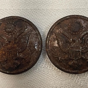 Pode incluir: Dois botões circulares antigos com um detalhado emblema de águia. Os botões são de um castanho escuro e desgastado, sugerindo idade e uso. A águia está centrada, com as asas abertas e um escudo por baixo.