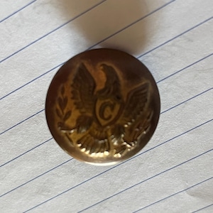 Puede incluir: Un botón dorado con un diseño de águila y la letra "C" en el centro.