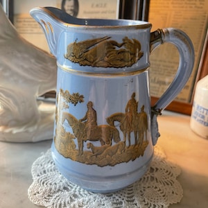 Puede incluir: Una jarra de cerámica azul y dorada con un diseño de escena de caza. La jarra tiene un asa y un pico. Está sentada sobre un encaje de ganchillo blanco.