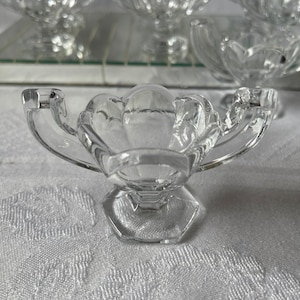 Puede incluir: Un plato de vidrio transparente con un borde festoneado y dos asas. El plato está sobre una superficie blanca.