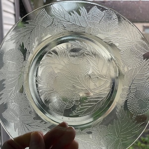 May include: A clear glass plate with an etched design of stylized thistle flowers and leaves. The plate has a raised rim and a smooth, glossy finish.