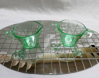 May include: Two green glass creamer or sugar bowls with handles. The bowls are on a silver mirrored surface.