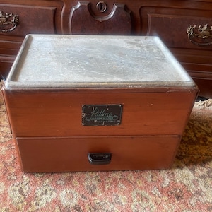May include: A vintage wooden box with a drawer and a hinged lid. The box is stained a warm brown color and has a metal nameplate on the front. The lid is a lighter color and appears to be made of a different material.