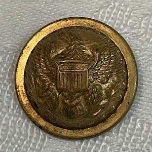 Puede incluir: Un botón de latón vintage con un diseño en relieve detallado. El botón presenta un emblema de escudo central, flanqueado por un águila con las alas extendidas y un borde exterior decorativo. El color del botón es latón cálido y envejecido.