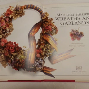May include: A book titled "Malcolm Hillier's Wreaths and Garlands" with a photograph of a wreath made with dried flowers and a ribbon. The wreath is a circular shape and is made with a variety of flowers in shades of pink, purple, and white.