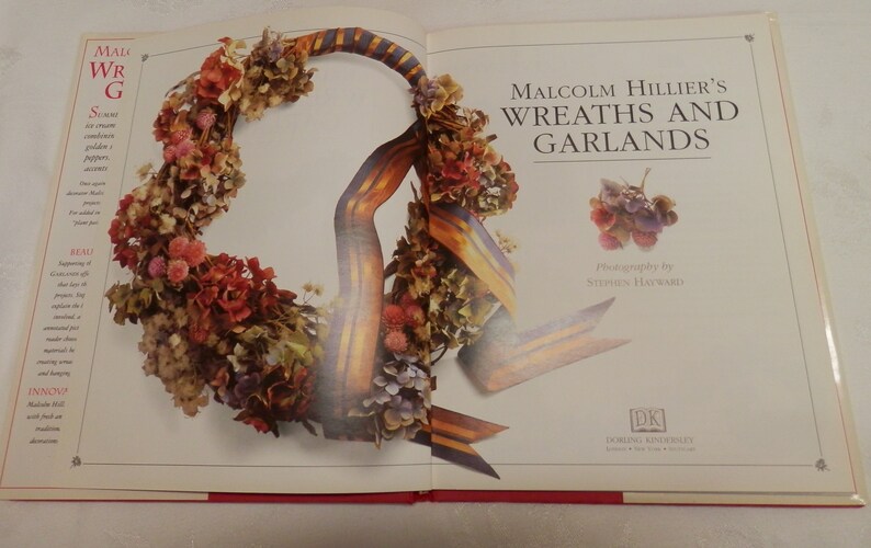 May include: A book titled "Malcolm Hillier's Wreaths and Garlands" with a photograph of a wreath made with dried flowers and a ribbon. The wreath is a circular shape and is made with a variety of flowers in shades of pink, purple, and white.