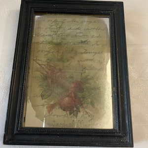 May include: A framed vintage postcard with a floral design and handwritten text. The text reads "A part of the price in July, but another Vision, the charm and the magic of the longest day." The bottom of the postcard reads "Gard of M."