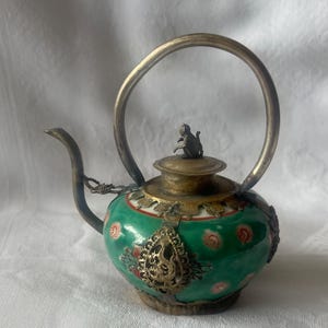 Könnte beinhalten: Eine kleine, grün-weiße Porzellan-Teekanne mit einem silbernen Henkel und Deckel. Die Teekanne ist mit Blumendesigns verziert und hat eine kleine, silberne Affenfigur auf dem Deckel.