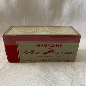Puede incluir: Una caja roja y blanca para un cambiador de diapositivas automático Airequipt. La caja está etiquetada "Magazine for Automatic Slide Changer" y "Holds 36 2" x 2" Slides".