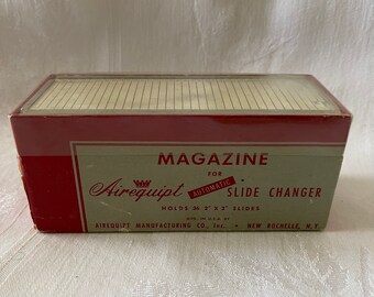 Può includere: Una scatola rossa e bianca per un cambia diapositive automatico Airequipt. La scatola è etichettata "Magazine for Automatic Slide Changer" e "Holds 36 2" x 2" Slides".