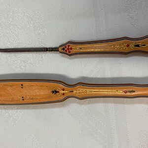 May include: Two antique crochet tools. The hook has a metal hook and a tan handle with gold and red accents. The other tool is a tan handle with gold and red accents.
