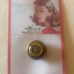 Puede incluir: Un botón vintage con un marco de metal dorado y un centro de vidrio transparente. El botón está en una tarjeta blanca con un borde rosa. La tarjeta tiene una imagen de una mujer con una flor en el pelo.