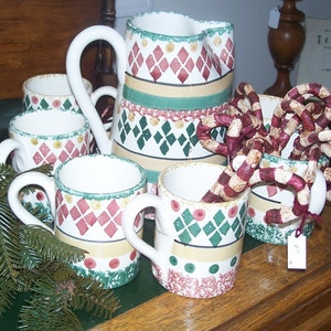 Puede incluir: Conjunto de cinco tazas de cerámica blanca con un patrón de diamantes verde, rojo y amarillo. Las tazas tienen un asa y están dispuestas sobre una superficie de madera con un fondo verde. Una jarra de cerámica blanca con un patrón similar también está incluida en el conjunto.