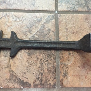 Könnte beinhalten: Ein dunkelgrauer Schraubenschlüssel aus Gusseisen mit einer quadratischen Öffnung an einem Ende und einem mehrzackigen Kopf am anderen. Das Werkzeug ist für einen bestimmten Zweck bestimmt, wahrscheinlich zum Anziehen oder Lösen von Muttern oder Bolzen.