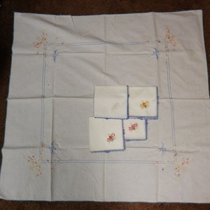 May include: A white tablecloth with blue embroidered edging and floral designs. Four white napkins with blue embroidered edging and floral designs are stacked in the center of the tablecloth.