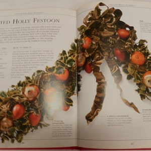 May include: A close-up of a fruited holly festoon, a Christmas decoration made with holly leaves, red berries, and apples. The festoon is tied together with a brown ribbon. The text "Fruited Holly Festoon" is visible in the image.