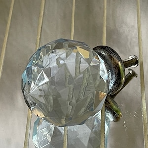 Puede incluir: Un pomo de cristal de vidrio transparente con una base de metal plateado y un tornillo. El pomo tiene un diseño facetado y es una pieza de ferretería decorativa.