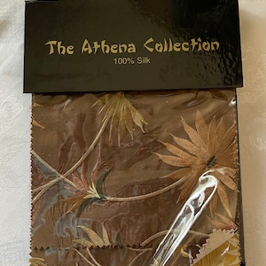 May include: A fabric swatch sample from The Athena Collection, featuring a brown background with a floral pattern in shades of gold, brown, and green. The label reads "The Athena Collection 100% Silk".