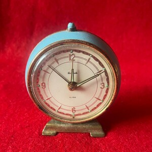 May include: A vintage blue alarm clock with a white face and gold accents. The clock has a simple design with black hands and a red alarm indicator. The clock face reads "ALARM" and has the text "H.L.CO."