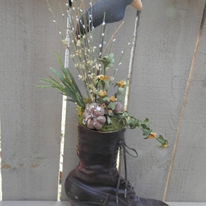 May include: A brown leather boot with a high heel is filled with a floral arrangement. The arrangement includes white and yellow flowers, green leaves, and a small gray bird.
