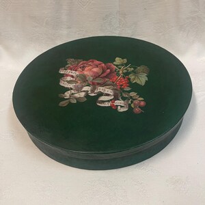 Puede incluir: Una caja redonda de color verde oscuro con un diseño floral en la tapa. El diseño presenta una gran rosa roja, bayas rojas y hojas verdes. También se incluye una pancarta con texto. La caja está sobre una superficie blanca.