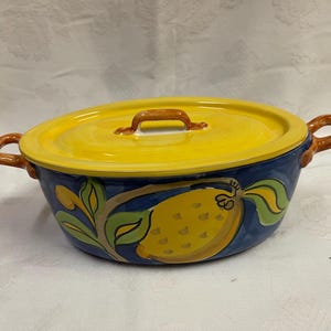 Puede incluir: Cazuela ovalada de cerámica con tapa y asas amarillas. El plato está pintado con una base azul y un diseño de limón. La tapa tiene un asa marrón. Las asas laterales también son marrones.