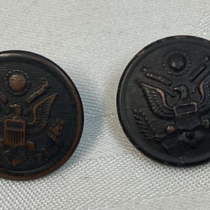 May include: Two antique bronze-coloured buttons featuring the Great Seal of the United States. Each button depicts an eagle with outstretched wings, a shield, and a banner. The buttons are round with a slightly raised design.