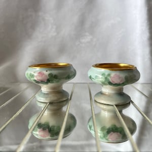 Könnte beinhalten: Zwei kleine, antike Porzellan-Sockelschalen. Die Schalen sind hellblau mit rosa Rosen- und grünen Blattmotiven. Die Ränder sind mit Gold verziert. Die Schalen spiegeln sich auf einer verspiegelten Oberfläche.