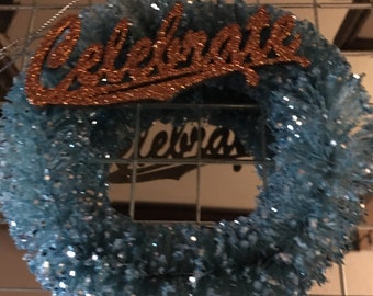 May include: A blue sparkly wreath with the word "Celebrate" in gold glitter letters.
