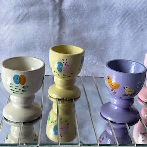 Könnte beinhalten: Fünf handbemalte Holz-Eierbecher mit verschiedenen Designs. Die Eierbecher sind in Blautönen, Gelb, Weiß, Rosa und Lila gehalten. Jeder Becher zeigt ein anderes Tierdesign, darunter ein Huhn, ein Vogel und ein Kaninchen.