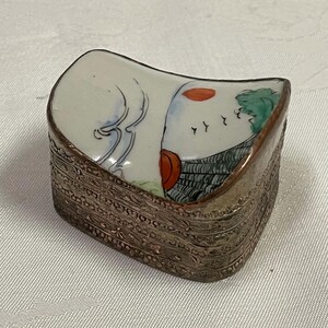 Puede incluir: Pequeña caja rectangular para baratijas con tapa curvada. La tapa presenta una escena pintada con una figura blanca, detalles en rojo y follaje verde. La caja tiene un marco de metal plateado ornamentado.